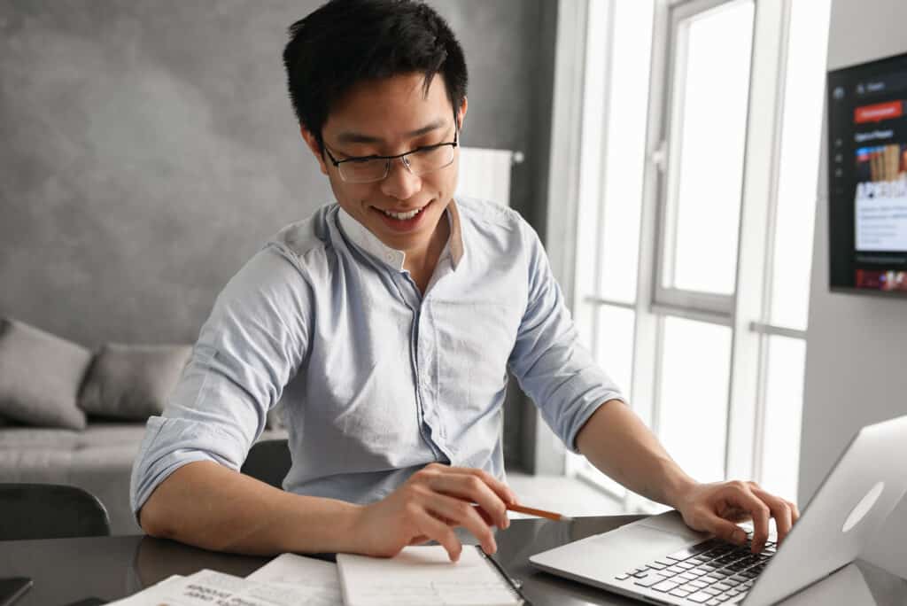 Portrait of a smiling young asian man using laptop computer while sitting at the table with paperwork at home